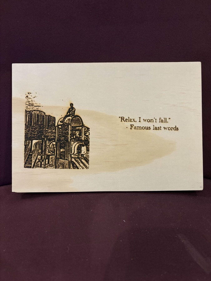 Engraved board of a person sitting on a rooftop with the quote 'Relax, I won't fall — famous last words'
