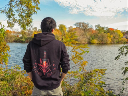 Person wearing a black Pyrolex hoodie with a red emblem, standing by a lake in autumn